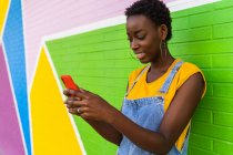 Afro-americano fêmea em macacão jeans em pé perto de parede colorida e celular de navegação — Fotografia de Stock