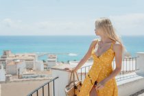 Женщина-путешественница в платье, стоящая возле забора и любуясь старым прибрежным городом и синим морем во время летних каникул — стоковое фото