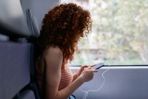 Mujer interesada con el pelo rizado en vaqueros rasgados mensajes de texto en el teléfono celular durante el viaje en tren durante el día - foto de stock