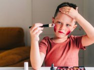 Очаровательный ребенок с аппликатором для макияжа трогает голову, глядя на стол с палитрой теней для век — стоковое фото