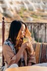 Donna etnica con lunghi capelli ondulati seduta sulla terrazza e sorseggiando cocktail rinfrescante da bicchiere con paglia mentre ammira il paesaggio in terrazza in Cappadocia, Turchia — Foto stock