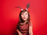 Joyful little girl in casual clothes and festive deer headband blowing kiss looking at camera during Christmas celebration against red background — Stock Photo