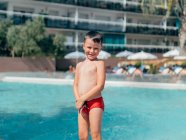 Positive shirtless boy in swimwear standing near swimming pool with clear rippling water in hotel during summer holidays — Stock Photo