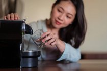 Jovem fêmea étnica contra máquina de café pod derramando bebida quente com espuma em vidro na cozinha da casa — Fotografia de Stock