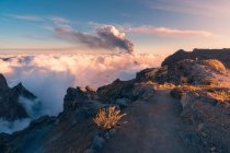 Nascer do sol em uma trilha de montanha de alta altitude em meio a nuvens brancas suaves e grossas e a erupção de um vulcão no fundo. Erupção vulcânica Cumbre Vieja nas Ilhas Canárias de La Palma, Espanha, 2021 — Fotografia de Stock