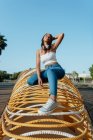 Cheerful young African American female in trendy wear squatting on symmetrical construction while looking away in city — Stock Photo