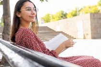 Positive young female in stylish clothes sitting with opened book on wooden bench against building with light wall in daytime — Stock Photo