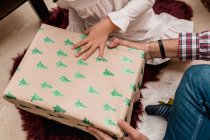 Обрезанный неузнаваемый ребенок, открывающий коробку подарков между анонимным отцом во время новогоднего праздника дома — стоковое фото