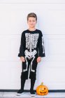 Corpo inteiro de menino pré-adolescente sorridente vestindo traje de Halloween preto com impressão de esqueleto em pé perto esculpida Jack O Lanterna abóbora contra a parede branca — Fotografia de Stock