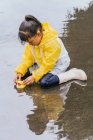 Очаровательный этнический ребенок в плаще играет с пластиковыми утками, отражающимися в волнистой луже в дождливую погоду — стоковое фото