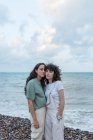 Junge lesbische Freundinnen in Freizeitkleidung umarmen sich, während sie an der Meeresküste unter bewölktem Himmel in die Kamera schauen — Stockfoto
