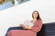 Mulher jovem positivo em roupas elegantes sentado com livro aberto no banco de madeira contra edifício com parede leve durante o dia — Fotografia de Stock