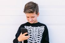 Веселий хлопчик в чорному скелеті костюм з пофарбованим обличчям, що використовується на мобільному телефоні, сидячи біля білої стіни на вулиці — стокове фото