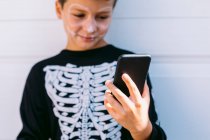Веселый мальчик в костюме черного скелета с раскрашенным лицом на мобильном телефоне, сидя возле белой стены на улице — стоковое фото