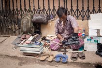 INDE, BANGLADESH - 6 DÉCEMBRE 2015 : Homme asiatique en vêtements traditionnels assis sur une clôture accroupi brillant des chaussures avec des marchandises diverses tout en travaillant sur le marché de la rue — Photo de stock