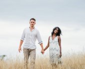 Homme avec petite amie ethnique tenant la main tout en marchant regardant la caméra sur la prairie d'automne sous un ciel nuageux — Photo de stock