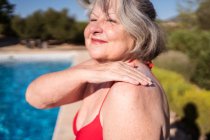 Vista lateral de una mujer despreocupada con pelo gris aplicando bloqueador solar en el hombro mientras disfruta de un día soleado junto a la piscina - foto de stock