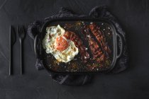 Vue du dessus de l'oeuf latéral ensoleillé avec tranches de bacon frit et condiments sur plateau contre des couverts sur fond sombre — Photo de stock