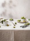 Натюрморт композиція з зелених свіжих слив, обладнана посудом на столі, покритим скатертиною — стокове фото