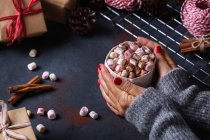 De cima da colheita caneca de retenção feminina de bebida quente com marshmallows entre cones de presentes de Natal e paus de canela — Fotografia de Stock