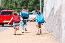 Visão traseira de escolares anônimos com mochilas correndo na passarela de azulejos na cidade ensolarada em fundo embaçado — Fotografia de Stock