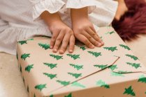 Récolter anonyme enfant ouverture boîte cadeau avec motif sapin sur le sol pendant les vacances du Nouvel An dans la maison — Photo de stock
