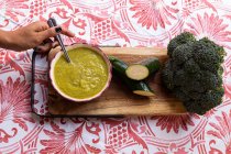 Top view of crop anonymous female with spoon in bowl with puree made of zucchini and broccoli on wooden chopping board — Stock Photo