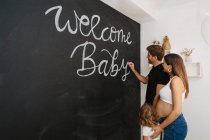 Male writing Welcome Baby inscription on blackboard against expectant female beloved and daughter in house — Stock Photo