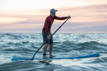 Vue latérale du surfeur masculin en combinaison et chapeau sur planche à pagaie surfant sur le bord de mer pendant le coucher du soleil — Photo de stock