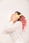 Vue latérale de la femelle calme aux cheveux roses en chemisier blanc debout les yeux fermés et écoutant de la musique dans les écouteurs sur fond de lumière — Photo de stock
