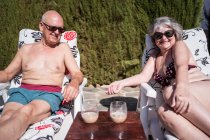 Casal sênior despreocupado em roupa de banho e óculos de sol banhos de sol em espreguiçadeiras com beber café perto da piscina e sebe — Fotografia de Stock