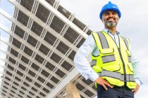 Desde abajo de sonriente contratista masculino étnico maduro en hardhat y chaleco de pie cerca de la estación de energía solar - foto de stock