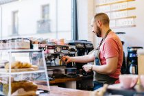 Vista laterale del barista maschio in grembiule con portafilter fermentazione caffè utilizzando macchina da caffè mentre in piedi vicino al bancone in caffetteria — Foto stock