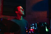 Концентрированный молодой музыкант играет на барабанах в клубе с зеленым и синим неоновым освещением — стоковое фото