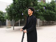 Trendy giovane ragazzo afroamericano con i capelli ricci scuri in abito elegante in piedi sulla piazza della città e guardando lontano dopo lo scooter di guida — Foto stock