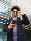 Bajo ángulo de alegre joven afroamericano masculino con pelo oscuro rizado en ropa de moda ajustando gafas de sol y sonriendo felizmente mientras está de pie cerca de un edificio moderno con teléfono inteligente en la mano - foto de stock