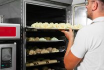 Jovem e elegante padeiro masculino em roupas casuais e óculos colocando croissant de massa crua em rack de metal enquanto assa pão na cozinha — Fotografia de Stock