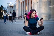 Full body of female photographer with pink hair and photo camera in hand surfing cellphone while sitting on walkway near aged building in city — Stock Photo