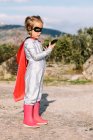 Боковой вид самоуверенной девушки в костюме супергероя с накидкой, просматривающей мобильный телефон — стоковое фото