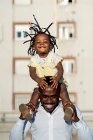 Alegre padre afroamericano en camisa llevando a su hijita en hombros y saltando mientras pasan tiempo juntos en la calle en la ciudad a la luz del sol - foto de stock
