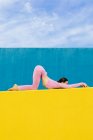 Vue latérale du corps entier jeune femme pratiquant la pose de yoga Uttana Shishosana tout en étant couché sur un mur jaune sur fond bleu — Photo de stock