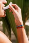 Side view of crop unrecognizable person with rainbow ribbon on hand biting slice of fresh natural coconut against green plants in summer time — Stock Photo