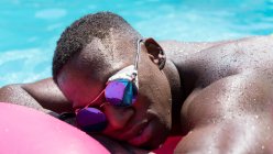 Homem afro-americano sem camisa em óculos de sol descansando sobre colchão inflável rosa na piscina enquanto toma sol no dia ensolarado de verão — Fotografia de Stock