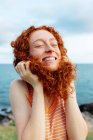 Joyful young redheaded female making childish pose with curly hair while enjoying freedom on seashore with eyes closed — Stock Photo