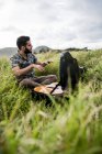 Side view of calm male musician in casual clothes sitting on green grass and opening black case of acoustic guitar on shore near sea in daylight — Stock Photo