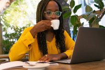 Afro-Américain boire du café chaud assis à la table en bois dans la cafétéria moderne tout en travaillant dans un ordinateur portable sur fond flou — Photo de stock