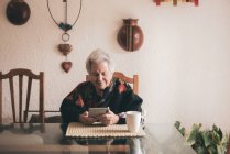 Smiling elderly female wearing warm clothes sitting at table with tablet and cup of tea looking at screen — Stock Photo