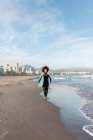 Jeune surfeuse réfléchie en combinaison avec planche de surf marchant en regardant loin sur le bord de mer lavé par la mer ondulante — Photo de stock