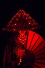 Mulher irreconhecível em roupa autêntica e chapéu tradicional com lâmpadas brilhantes em pé no estúdio escuro com ventilador na mão sobre fundo preto — Fotografia de Stock