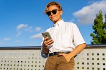 Снизу молодой мужчина в белой рубашке пишет смс по мобильному телефону, стоя на улице против голубого неба в солнечный день — стоковое фото